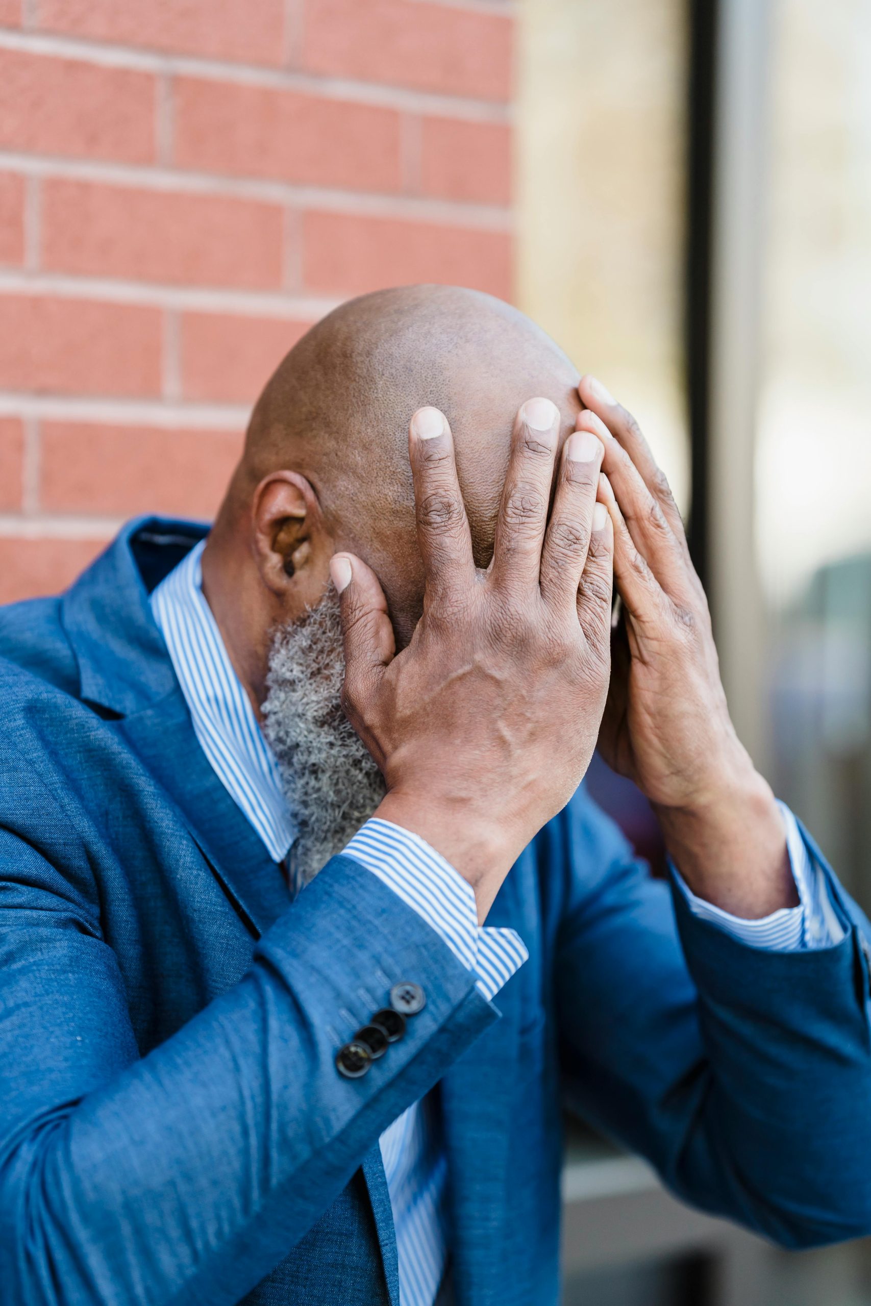Middle-aged man in a blue suit looking stressed with hands on face outdoors.
